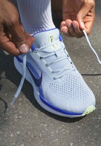 Zapatilla deportiva gris claro con acentos azules, parte superior de malla y cordones blancos. La suela presenta un patrón texturizado. Persona atándose los cordones al aire libre.