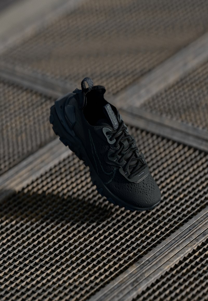 Chaussure de sport noire avec semelle texturée flottant au-dessus d'une surface en grille métallique dans une lumière naturelle douce.
