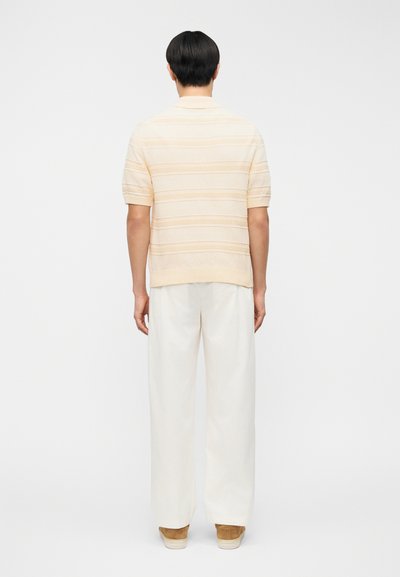 Κοντομάνικο, ελαφρύ μπεζ πλεκτό πόλο με υφασμάτινες ρίγες, φορεμένο με λευκό παντελόνι φαρδιάς γραμμής και καφέ παπούτσια. Ορατό λείο ύφασμα και γιακάς.