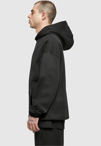 Felpa nera oversize con tasca a marsupio, materiale spesso e cappuccio ampio. Presenta cuciture sottili e polsini a coste.