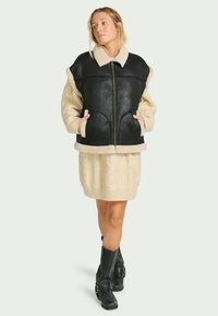 Casaco oversized preto e creme com fecho por zip, mangas texturizadas e punhos e bainha canelados. Usado sobre um vestido creme de malha em ponto de cable.