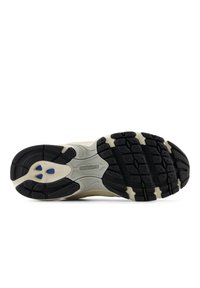 Sola de borracha preta com um padrão texturizado, apresentando uma entressola off-white e um detalhe azul. Logótipo da New Balance posicionado centralmente.