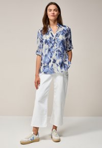 Camisa ligera y estampada en azul y blanco, con cuello y mangas enrolladas, combinada con pantalones anchos blancos y zapatillas beige.