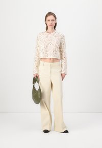 DickerStrick, cropped Cremepullover mit strukturiertem Muster; hochgeschnittene beiger weit geschnittene Hose; grüne Schultertasche; schwarze flache Schuhe.