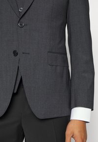 Mörkgrå kavaj med texturerad tyg, enkelknäppt design, tre knappar och bröstficka. Vitt skjortärm synlig.