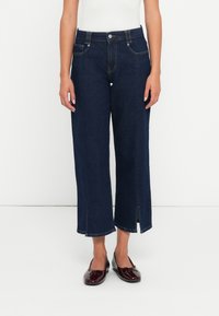 Jeans largi de culoare albastră închisă, din denim, având buzunare frontale, fermoar la brâu și slituri la tiv. Asortați cu pantofi negri fără toc.