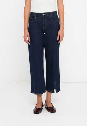 Mørkeblå vide jeans laget av denim, med frontlommer, glidelås og splitt i kanten. Sammen med svarte flate sko.