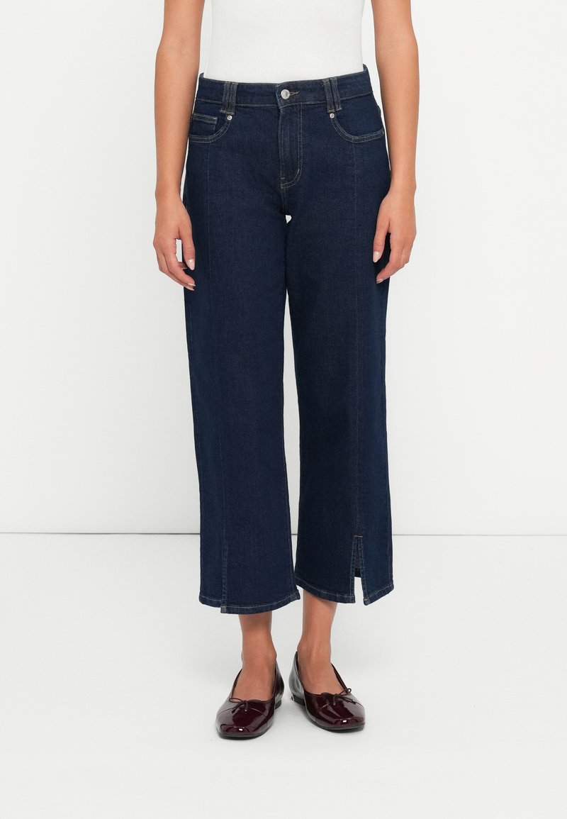 Mørkeblå vide jeans laget av denim, med frontlommer, glidelås og splitt i kanten. Sammen med svarte flate sko.