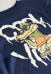 Navyblaue Baumwoll-T-Shirt mit einem verspielten Cartoon-Krokodil-Graphic, das Sonnenbrillen und ein grünes Outfit trägt, vor einem weißen Pinselstrich-Hintergrund.