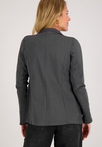 Grijze blazer met lange mouwen, een enkele kraag, getailleerde pasvorm en zichtbaar stiksel. Glad materiaal met een iets textuur gegeven oppervlak.