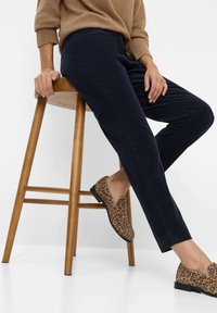 Navyblauwe corduroy broek gecombineerd met laarzen met luipaardprint. Het model zit op een houten kruk en draagt een tan kleurige sweater.