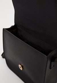 Sort læderhåndtaske med en glat overflade, prydet med en gyldent cirkulær lås og et enkelt design. Indersiden er uforet og mørk.