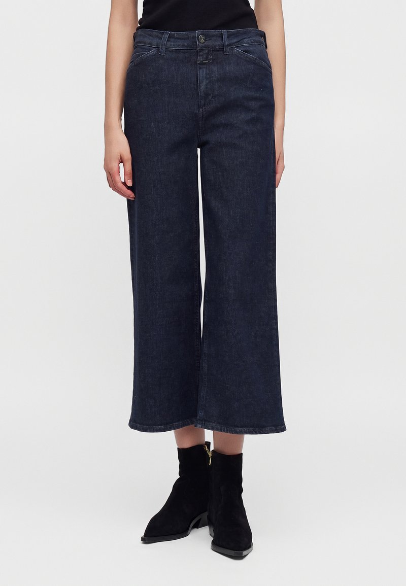 Γυναίκα που φοράει σκούρα μπλε, cropped wide-leg τζιν και μαύρα μποτάκια αστραγάλου, στέκεται μπροστά σε απλό φόντο.