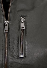 Schwarze Lederjacke mit zwei Reißverschlusstaschen, silberfarbener Hardware und feiner Oberflächenstruktur mit detaillierter Steppnaht entlang der Kanten.