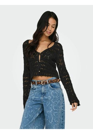 Ženska, oblečena v črno kvačkano kratko majico z dolgimi rokavi, modre kavbojke z visokim pasom in vzorcem ter pas z leopardjim potiskom, pozira z zaprtimi očmi.