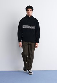 Czarny polarowy pulower z pół-zamkiem, z szarym logo Quiksilver na piersi, połączony z oliwkowymi spodniami i czarnymi trampkami.