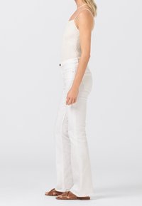 Białe dżinsy w kształcie dzwonów wykonane z denimu, w połączeniu z prążkowanym jasnobeżowym topem na ramiączkach. Modelka stoi bokiem, w brązowych sandałach na paski.