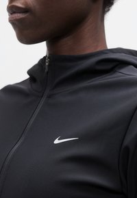 Κοντινό πλάνο ενός ατόμου που φοράει μαύρο μπουφάν με φερμουάρ και λευκό λογότυπο Nike στο στήθος, στραμμένο προς τα αριστερά.