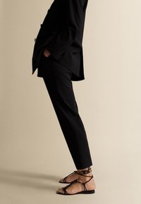 Musta räätälöity bleiseri yhdistettynä mustiin housuihin ja nauhakenkiin. Huoliteltu kangas, jäsennelty muotoilu ja minimalistiset yksityiskohdat näkyvät.