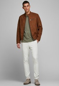 Brun faux suede jakke, olivengrøn t-shirt, hvide slim-fit bukser og brune sko. Jakken har lynlås i siden og opretstående krave.