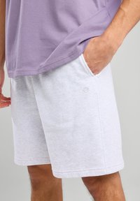 Vaaleanharmaat puuvillahortsit, joissa on teksturoitu pinta, sivutaskut ja pieni brodeerattu logo. Yhdistetty laventelinvärisen T-paidan kanssa.