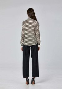 Femme aux longs cheveux sombres portant un chemisier beige à manches longues à motifs, un pantalon noir à larges jambes et des escarpins noirs, de dos.