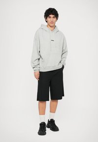 Szary hoodie z kieszenią z przodu i czarnym logo, w połączeniu z luźnymi czarnymi szortami. Czarne sneakersy i białe skarpety dopełniają wygląd.