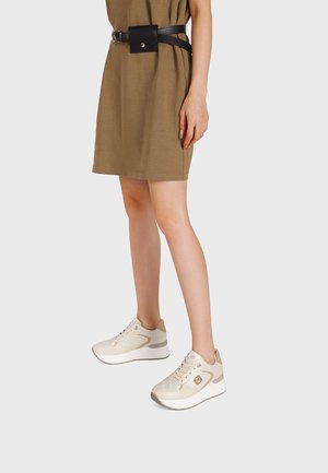Donna con sneakers beige, un vestito corto marrone e una cintura nera con borsa, su uno sfondo chiaro uniforme.