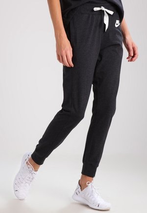 Person iført mørkegrå Nike træningsbukser med hvid snor og hvide vævede sneakers, stående på ét ben mod en ensfarvet baggrund.