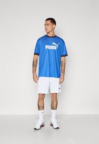 Blå atletisk skjorte med lodrette striber, prydet med et stort hvidt Puma-logo, parret med hvide shorts og hvide sko med røde accenter.