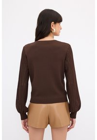 Brauner Strickpullover mit langen, strukturierten Ärmeln. Tanfarbene Shorts sind darunter sichtbar. Der Pullover hat einen gerippten Saum und ein offenes Muster an den Ärmeln.