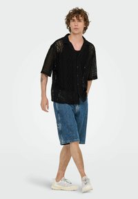 Camisa negra de punto de manga corta con una textura de patrón, combinada con unos pantalones cortos de mezclilla azules y zapatillas de color claro. Modelo en una pose caminando.