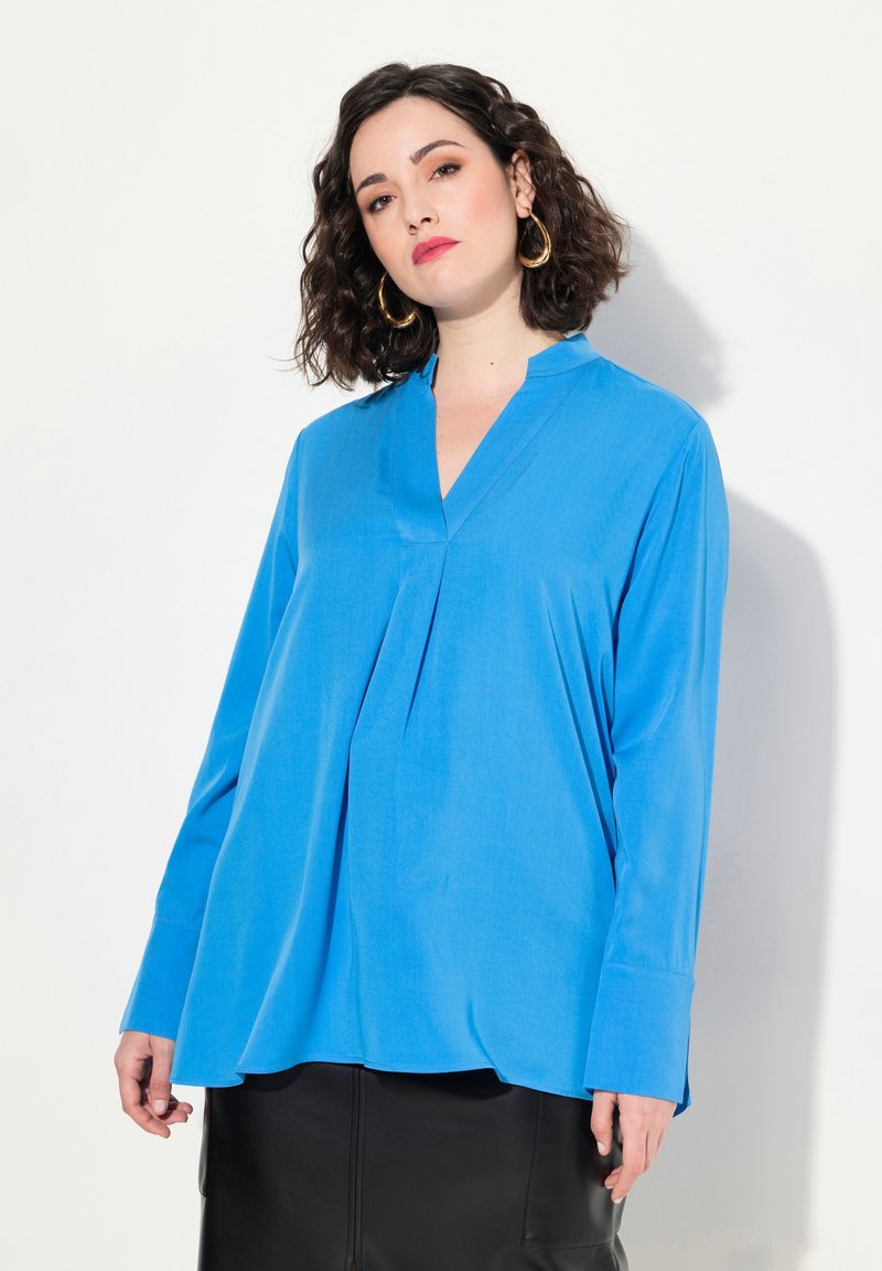 Blusa blu a maniche lunghe con colletto a V, caratterizzata da una piega in tessuto morbido. Abbinata a una gonna di pelle nera.