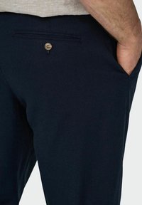 Pantaloni blu navy con una tasca frontale e una tasca posteriore chiusa con un bottone in ottone. Tessuto dalla superficie liscia e design su misura.