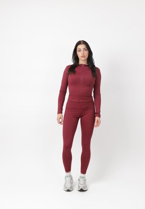 Donna in piedi rivolta frontalmente, indossa una giacca aderente color bordeaux con cerniera e leggings coordinati, con scarpe da ginnastica bianche, su uno sfondo bianco uniforme.
