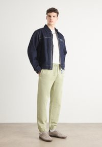 Jeansjacke mit kontrastierenden Nähten, weißes T-Shirt, hellgrüne, schmal zulaufende Hose, graue Sneaker mit Schnürsenkeln und einem Akzent-Reißverschlussdetail.