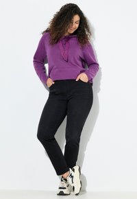 Sudadera con capucha morada, con cordón y bolsillo frontal, combinada con jeans negros que tienen detalles laterales y zapatillas blancas.