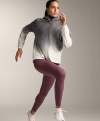 Ľahká, kapucňová bunda v gradientnej šedej a bielej farbe, skombinovaná s priliehavými fialovými legínami. Pozícia pri behu, viditeľná športová obuv.