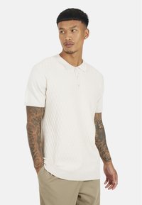 Koszulka polo z krótkim rękawem w jasnym beżu z teksturowanym wzorem w romby, z prążkowanym kołnierzykiem i mankietami, z trzema guzikami.