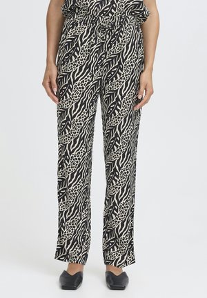 Mujer con pantalones sueltos en negro y beige con estampado abstracto de animal y zapatos planos negros con punta puntiaguda, de pie sobre un fondo liso.