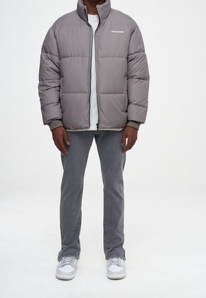 Osoba v šedé péřové bundě, bílém tričku, šedých džínách a bílých teniskách stojící před jednoduchým bílým pozadím.