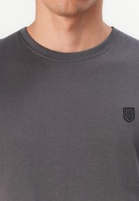 Graues Baumwoll-T-Shirt mit Rundhalsausschnitt, das auf der linken Seite der Brust ein kleines schwarzes Wappenlogo trägt. Weiche Textur, minimalistisches Design.