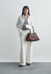 Fonott sötétbarna bőr kézitáska két füllel, fehér bőszárú nadrágot, szürke felsőt és fehér gombos inget viselő modell mellett bemutatva.