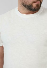 Weißes kurzärmliges T-Shirt aus leichtem Stoff, mit Rundhalsausschnitt und subtiler Textur. Kleines Logo-Detail auf der Brust.