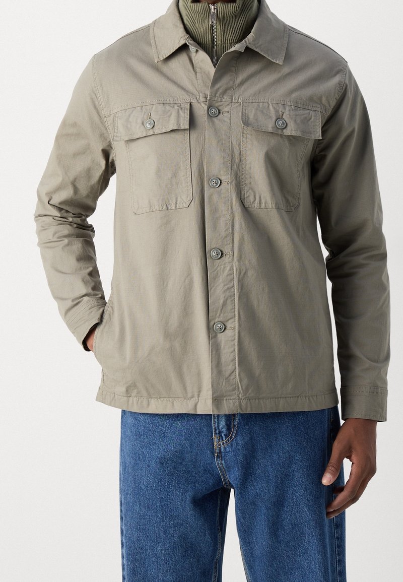 Homme portant une veste beige boutonnée avec deux poches poitrine sur un pull vert zippé et un jean bleu, une main dans la poche.