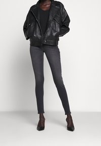 Chaqueta de biker de cuero negro con paneles texturizados, diseño en capas; jeans ajustados gris oscuro; tacones marrones con acentos negros en la punta.