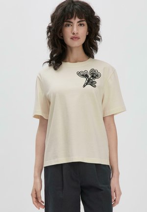 Crème-kleurig t-shirt met korte mouwen, voorzien van een zwart geborduurd bloemenmotief op de linkerborst. Zachte textuur met een casual pasvorm.