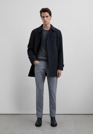 Abrigo oscuro de lana con doble botonadura sobre un suéter gris, combinado con pantalones gris claro y botines de cuero negro. Diseño sencillo y colores neutros.