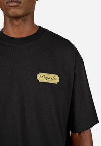 Melna kokvilnas T-kreklu ar īsām piedurknēm, ar zeltītu taisnstūrveida logo uzlīmi, kurā rakstīts "Pegador", kreisajā krūšu daļā.