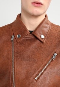 Giacca da motociclista in pelle marrone con finitura testurizzata, colletto a scatto e due tasche con zip dotate di dettagli in metallo argento.